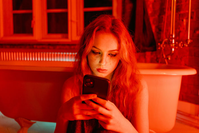 A woman sitting by a bathtub, focused on her phone, in a dimly lit room with red lighting. A woman sitting by a bathtub, focused on her phone, in a dimly lit room with red lighting.