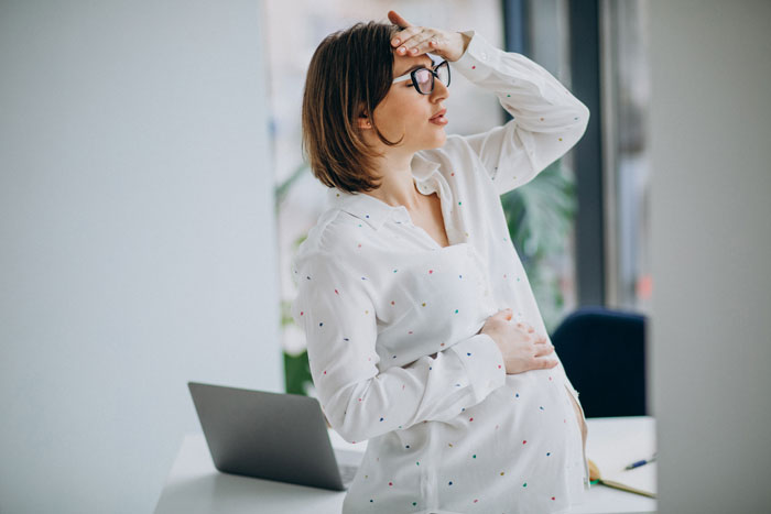 Pregnant woman in white shirt, holding her head, standing near a laptop, appearing concerned. Pregnant woman in white shirt, holding her head, standing near a laptop, appearing concerned.