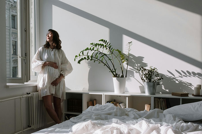 Pregnant woman in a serene bedroom, standing by the window with light streaming in and plants in the background. Pregnant woman in a serene bedroom, standing by the window with light streaming in and plants in the background.