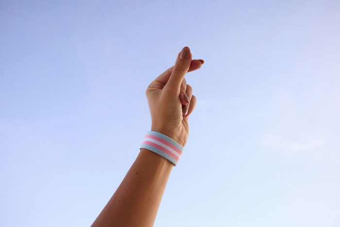 A hand with a pink and blue wristband raised against a clear sky, symbolizing transgender pride and identity. A hand with a pink and blue wristband raised against a clear sky, symbolizing transgender pride and identity.