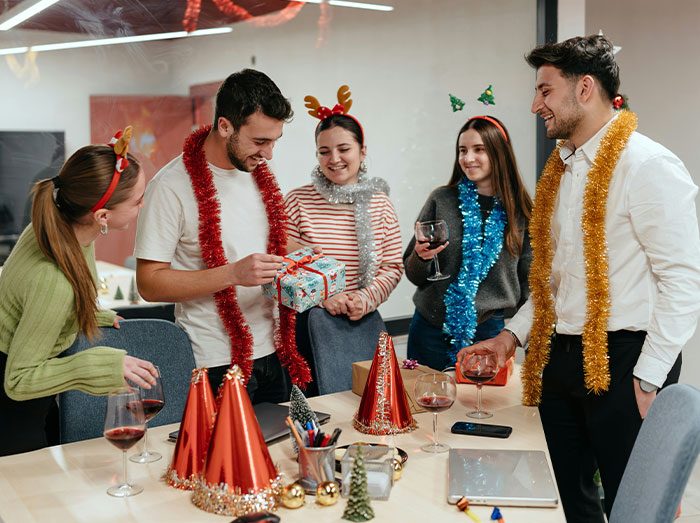 Office Christmas party with people in festive accessories, exchanging gifts and holding drinks around a decorated table. Office Christmas party with people in festive accessories, exchanging gifts and holding drinks around a decorated table.
