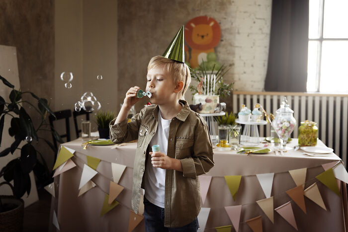 Boy wearing a party hat blows bubbles near a decorated table during a festive event. Boy wearing a party hat blows bubbles near a decorated table during a festive event.