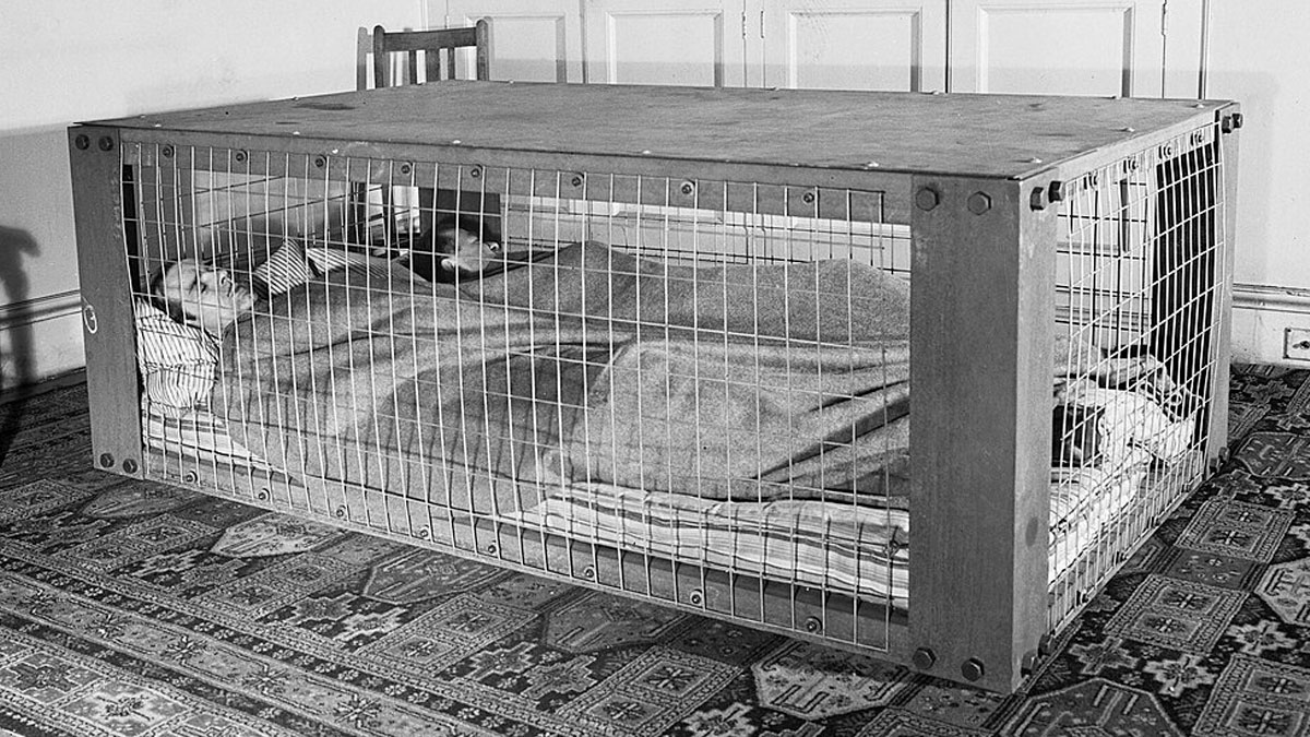 Man lying inside a strange invention cage bed covered with a blanket on a patterned floor, vintage black and white photo.