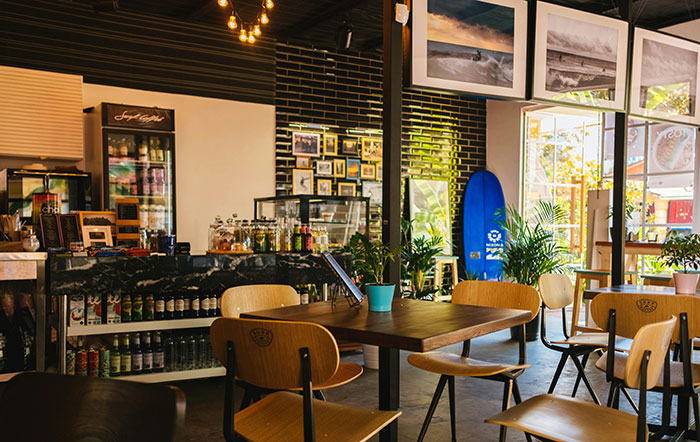 Cozy cafe interior with wooden chairs, small tables, and a surfboard by the window, evoking a trendy and relaxed atmosphere. Cozy cafe interior with wooden chairs, small tables, and a surfboard by the window, evoking a trendy and relaxed atmosphere.