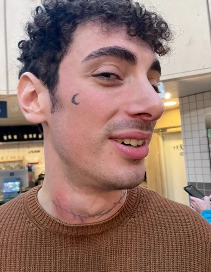 Person with a crescent moon temporary tattoo on cheek and faded tattoo on neck, wearing a brown sweater indoors. Person with a crescent moon temporary tattoo on cheek and faded tattoo on neck, wearing a brown sweater indoors.