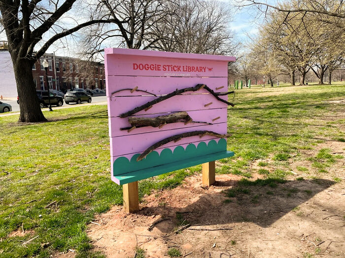 A pink wooden stick library for dogs stands in a park, filled with branches and labeled "Doggie Stick Library. A pink wooden stick library for dogs stands in a park, filled with branches and labeled "Doggie Stick Library.