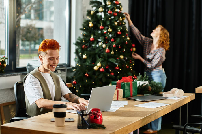 Woman decorating Christmas tree, focusing on Secret Santa planning, with a man using a laptop in the office. Woman decorating Christmas tree, focusing on Secret Santa planning, with a man using a laptop in the office.