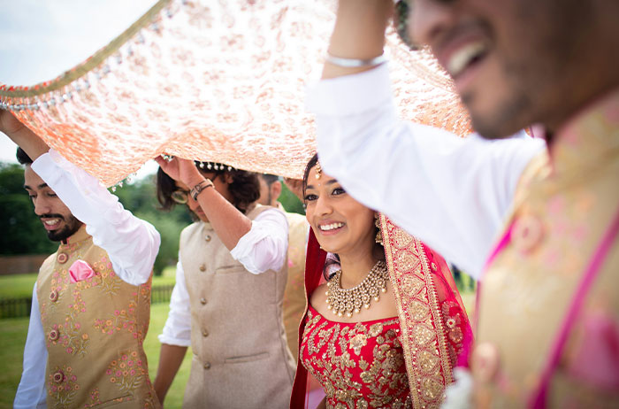 Bride in traditional attire smiling joyfully under ornate canopy at an outdoor wedding ceremony, surrounded by guests. Bride in traditional attire smiling joyfully under ornate canopy at an outdoor wedding ceremony, surrounded by guests.