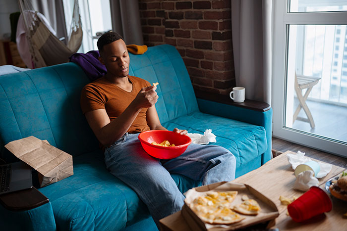 Man in an orange shirt sits on a blue couch, eating from a red bowl, highlighting roommate dynamic issues. Man in an orange shirt sits on a blue couch, eating from a red bowl, highlighting roommate dynamic issues.