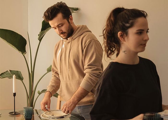 Man in beige hoodie setting table, while woman in black shirt looks away, emphasizing relationship boundaries. Man in beige hoodie setting table, while woman in black shirt looks away, emphasizing relationship boundaries.
