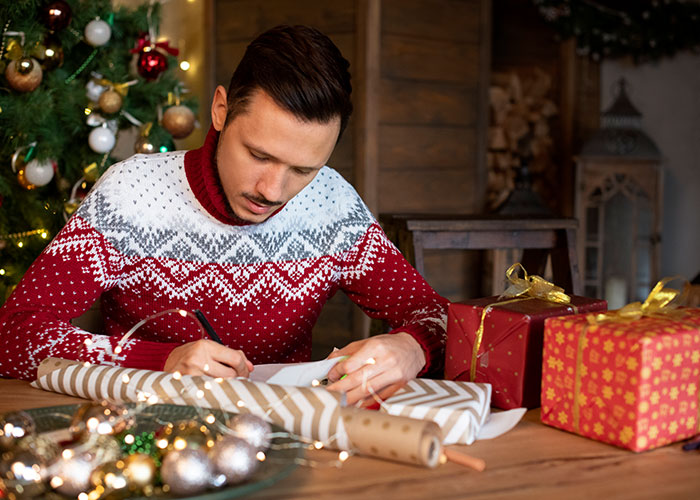 Man wrapping Christmas gifts at a wooden table, surrounded by festive decorations, focusing on holiday preparations. Man wrapping Christmas gifts at a wooden table, surrounded by festive decorations, focusing on holiday preparations.