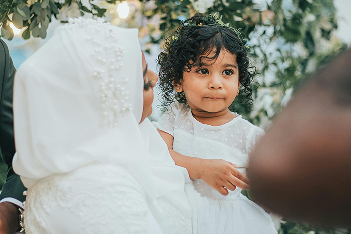 Child at a wedding, surrounded by greenery, holding onto an adult, illustrating family dynamics at events. Child at a wedding, surrounded by greenery, holding onto an adult, illustrating family dynamics at events.