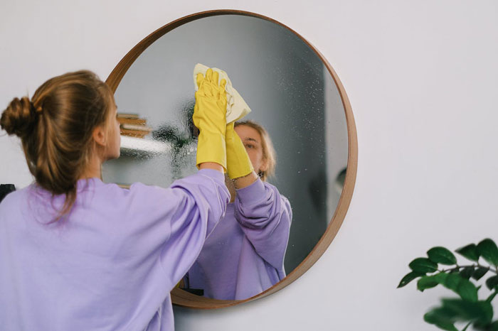 Woman in yellow gloves cleaning a mirror, reflecting a tidy room. Woman in yellow gloves cleaning a mirror, reflecting a tidy room.
