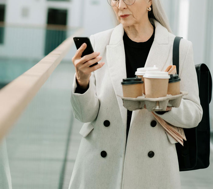 An older woman with takeout coffee checks her phone, illustrating unexpected visit dynamics. An older woman with takeout coffee checks her phone, illustrating unexpected visit dynamics.