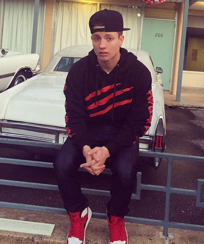 Young Matt Rife in a black hoodie and red shoes sitting on a fence in front of a vintage car Young Matt Rife in a black hoodie and red shoes sitting on a fence in front of a vintage car