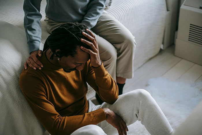 A worried man in a brown sweater sits on the floor, comforted by a person beside him, symbolizing tension over a birthday incident. A worried man in a brown sweater sits on the floor, comforted by a person beside him, symbolizing tension over a birthday incident.