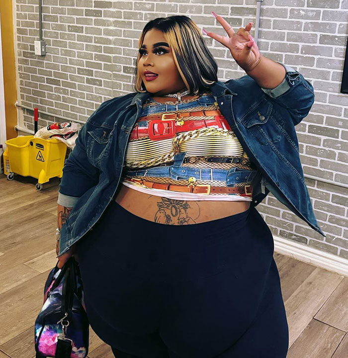 Plus size woman in colorful top and denim jacket poses with a peace sign in a brick-walled room. Plus size woman in colorful top and denim jacket poses with a peace sign in a brick-walled room.