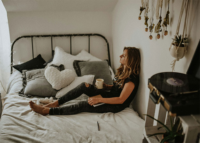 Woman sitting on a bed with a mug, surrounded by cozy cushions and decor, relating to jokes about stains in her home. Woman sitting on a bed with a mug, surrounded by cozy cushions and decor, relating to jokes about stains in her home.