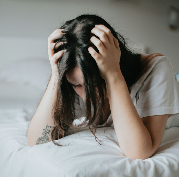 Woman stressed, holding her head, on a bed, pondering financial strain from not buying a friend's replacement hairdryer. Woman stressed, holding her head, on a bed, pondering financial strain from not buying a friend's replacement hairdryer.