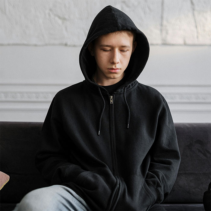 Teen in a black hoodie sitting on a sofa, looking down and appearing contemplative. Teen in a black hoodie sitting on a sofa, looking down and appearing contemplative.