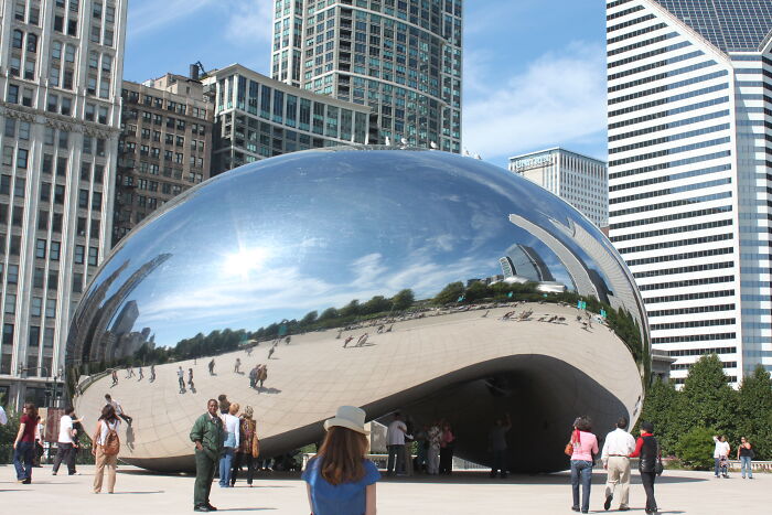 People admire the reflective sculpture in a park, surrounded by tall buildings, under a clear sky, showcasing amazing views.