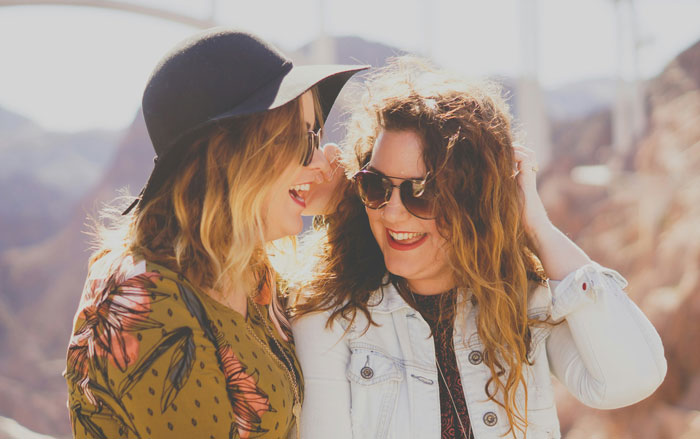Two women smiling in sunlight, wearing sunglasses and casual outdoor clothing, enjoying a moment together. Two women smiling in sunlight, wearing sunglasses and casual outdoor clothing, enjoying a moment together.
