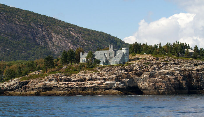 Coastal mansion with stunning views, situated on a rocky shoreline against a backdrop of mountains and blue sky.