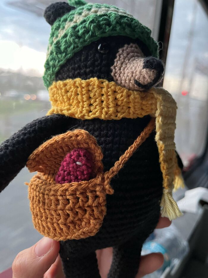 Crochet bear doll wearing a green hat and yellow scarf, holding a crocheted basket with colorful items inside.