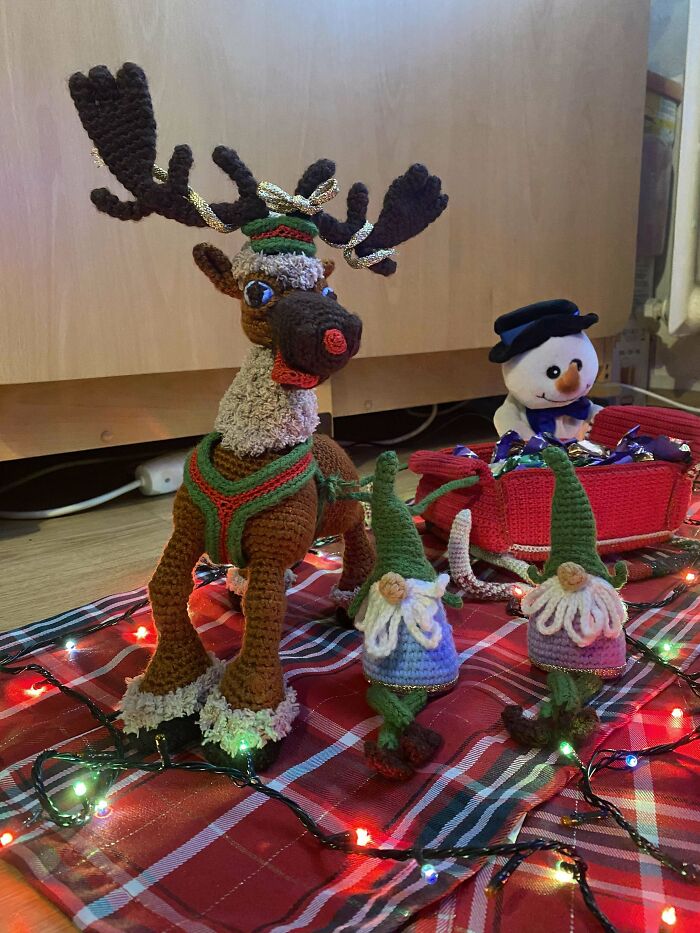 Crochet reindeer and gnome figures with a snowman in a festive holiday setting with colorful string lights.