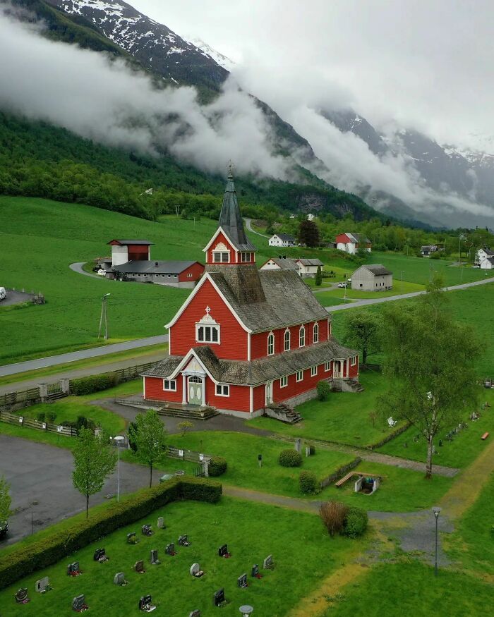 Red church in a lush valley with mountains and gravestones, showcasing fascinating architecture and scenic beauty.