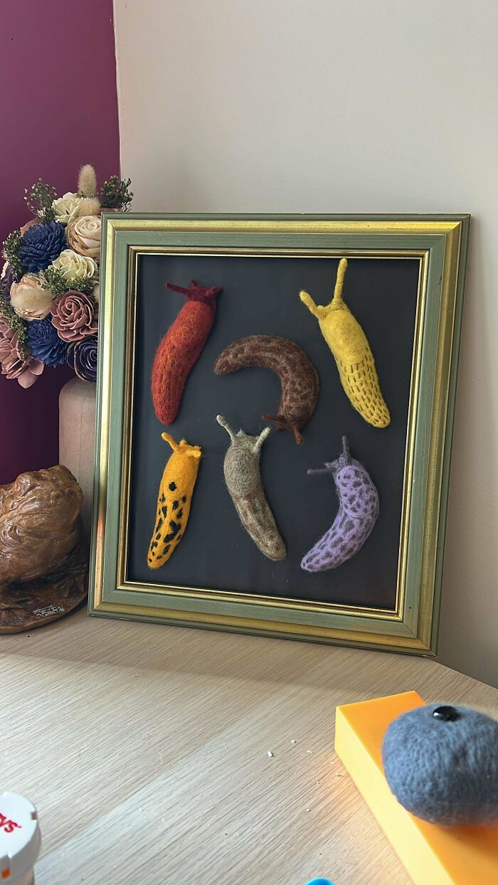 Framed DIY projects featuring colorful felted slugs displayed on a table with flowers and a clay sculpture.