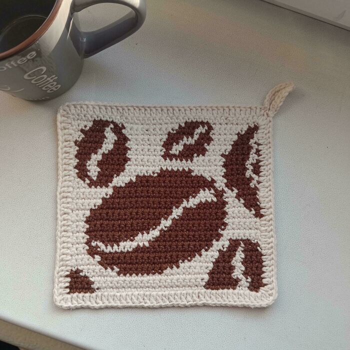 Crochet enthusiasts proudly display a handmade coffee bean patterned crochet square with a hanging loop next to a coffee mug.