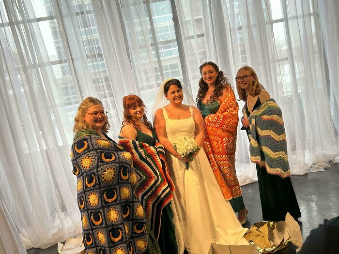 Five women, one in a wedding dress, proudly showcase colorful crochet blankets at a celebration event indoors.