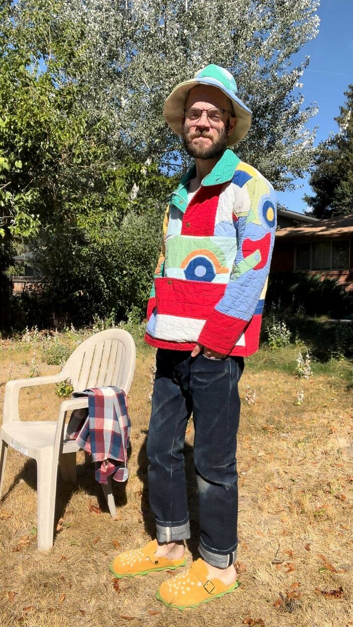 Man in colorful DIY quilted jacket, wearing a bucket hat and bright shoes, stands in a sunny garden next to a chair.