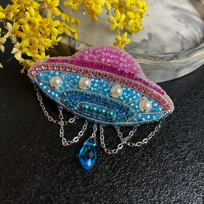 Beaded UFO brooch DIY project with chains and blue gem accent, set on black surface with yellow flowers in the background.