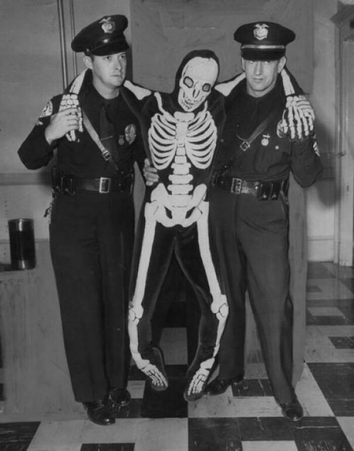 Two officers holding a person in a skeleton costume in a vintage black and white photograph.
