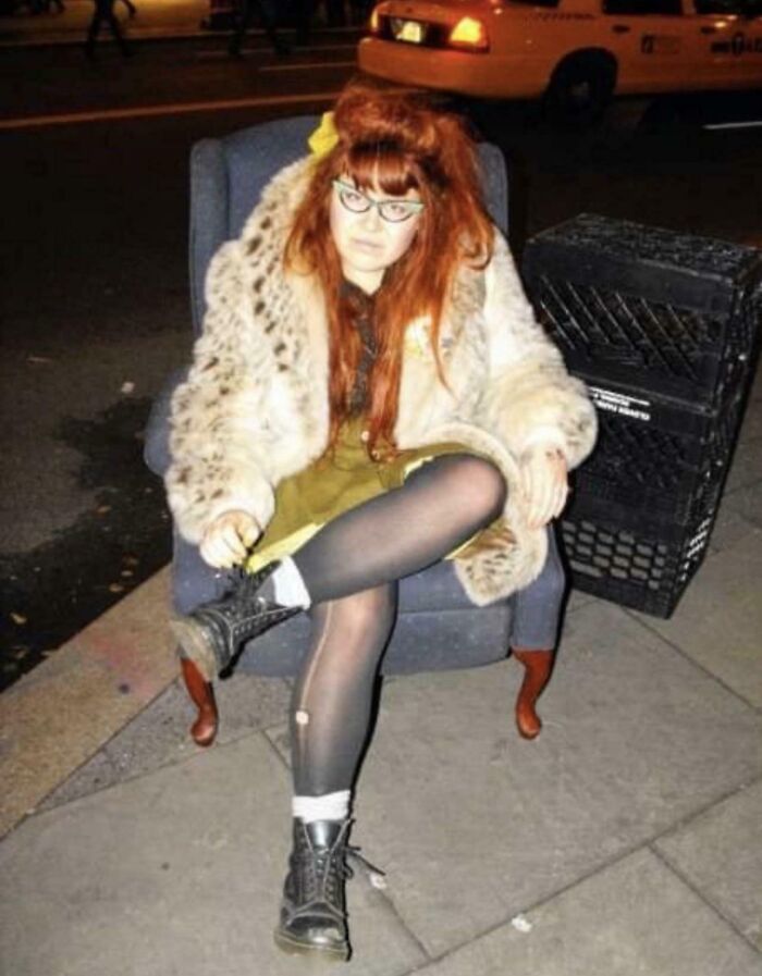 Person in vintage attire sitting on a street chair, embodying blunder years style with a fur coat and glasses.
