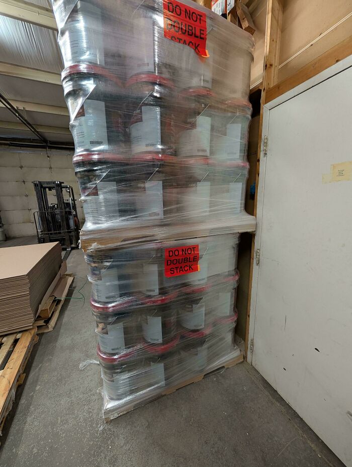 Stacked pallets with containers wrapped in plastic disregarding safety protocol signs in an industrial warehouse setting.