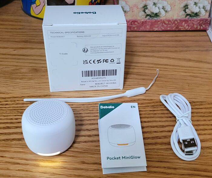 Travel essentials: compact speaker with packaging, manual, lanyard, and USB cable on a wooden desk.