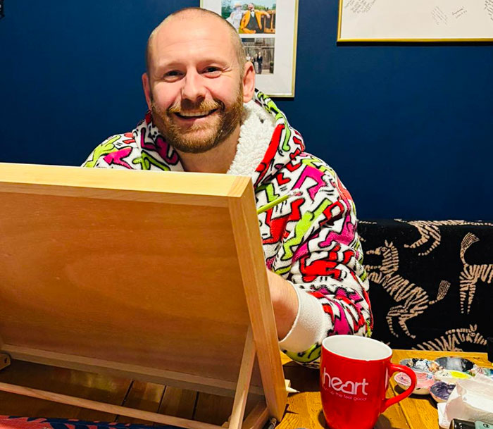 Man smiling while painting, wearing a colorful hoodie, with a red mug labeled "heart" nearby, related to viral painting. Man smiling while painting, wearing a colorful hoodie, with a red mug labeled "heart" nearby, related to viral painting.
