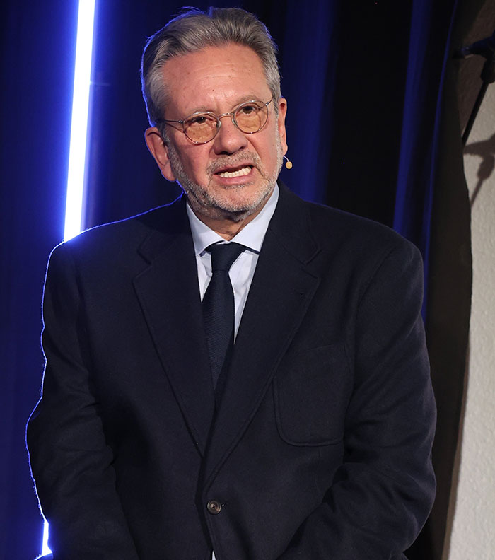 Man in a suit with glasses speaking on stage about elegance and class. Man in a suit with glasses speaking on stage about elegance and class.