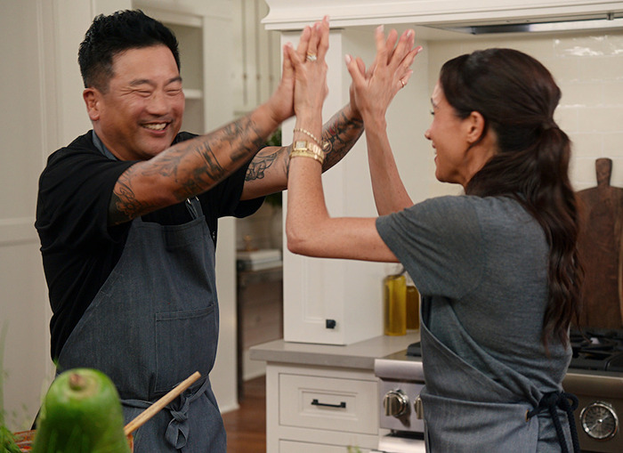 Meghan Markle high-fiving guest in a kitchen, wearing aprons. Meghan Markle high-fiving guest in a kitchen, wearing aprons.