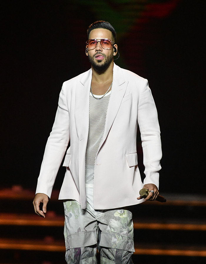 Romeo Santos on stage in a white jacket and floral pants, holding a microphone. Romeo Santos on stage in a white jacket and floral pants, holding a microphone.