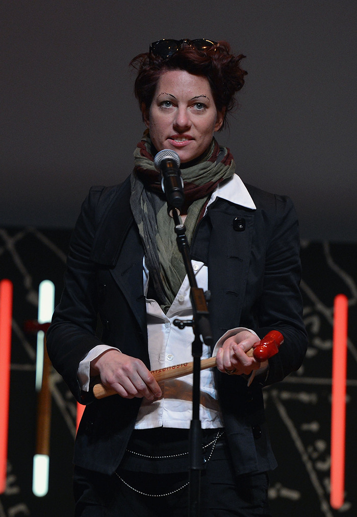Amanda Palmer in a black jacket and scarf holding a microphone on stage. Amanda Palmer in a black jacket and scarf holding a microphone on stage.