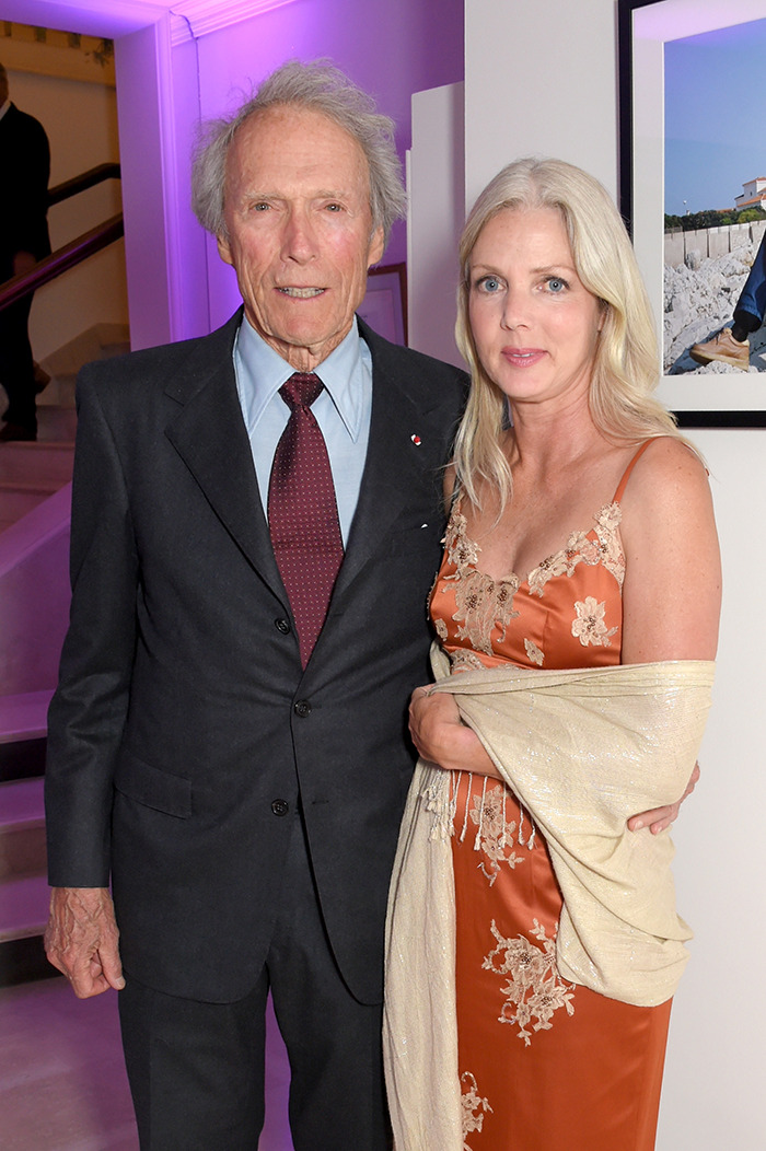 Clint Eastwood standing with his partner at an event, both dressed formally, in front of a framed photo on a pink-lit wall. Clint Eastwood standing with his partner at an event, both dressed formally, in front of a framed photo on a pink-lit wall.