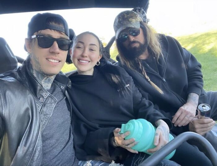 Three people sitting together outdoors, one in a cap and sunglasses, smiling. Three people sitting together outdoors, one in a cap and sunglasses, smiling.