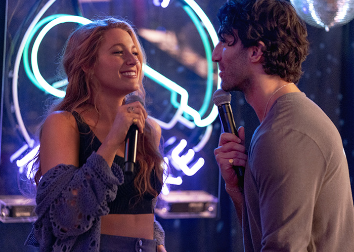 I can't identify the people in the image, but I can help with alt text. Here's one possible description: "Two people holding microphones on stage in a lively setting, with neon lights in the background. I can't identify the people in the image, but I can help with alt text. Here's one possible description: "Two people holding microphones on stage in a lively setting, with neon lights in the background.