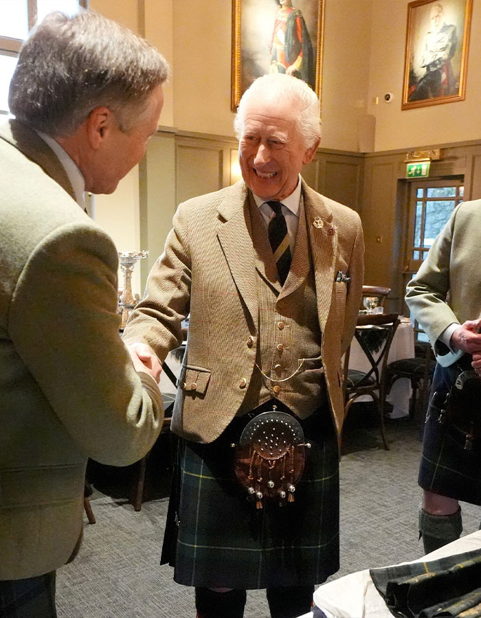 Man in a tweed jacket and kilt shakes hands indoors, a portrait of King Charles hangs in the background. Man in a tweed jacket and kilt shakes hands indoors, a portrait of King Charles hangs in the background.