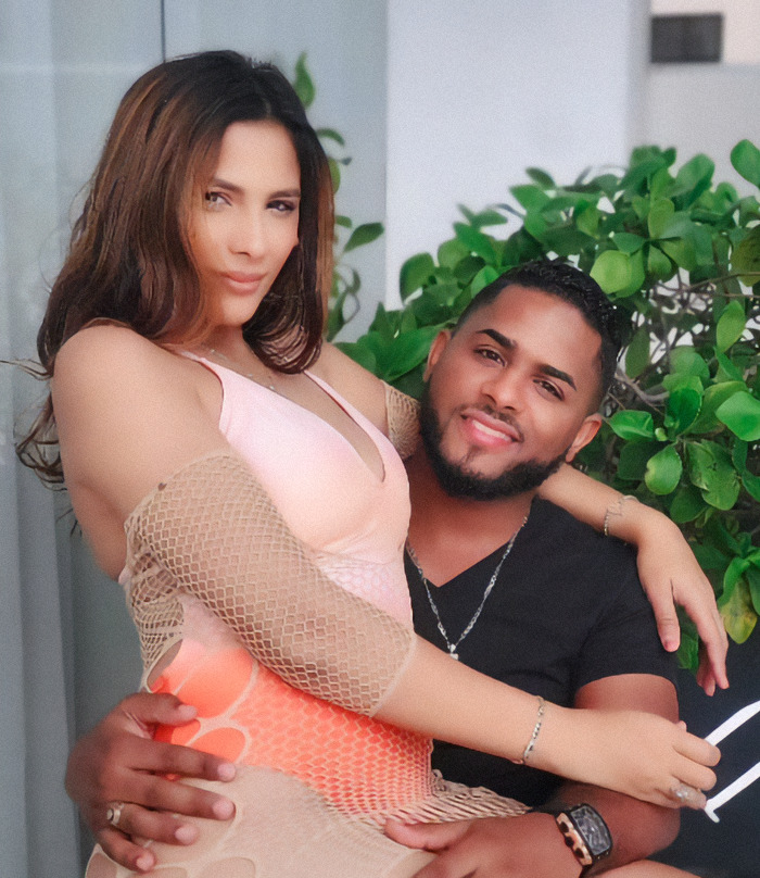 Couple sitting together, woman in a peach dress and man in a black shirt, smiling with greenery in the background. Couple sitting together, woman in a peach dress and man in a black shirt, smiling with greenery in the background.