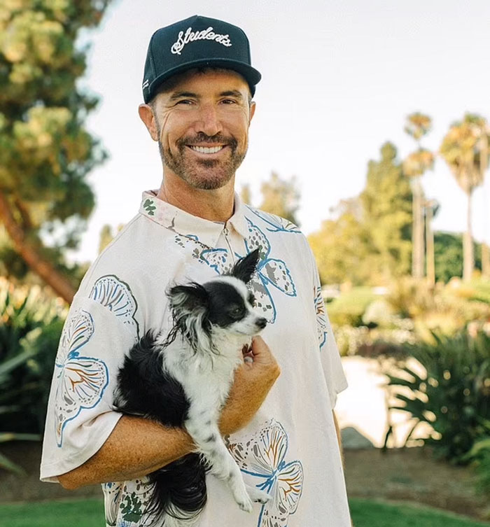 Man in a floral shirt and cap holding a small dog in a sunny garden setting, related to MySpace founder. Man in a floral shirt and cap holding a small dog in a sunny garden setting, related to MySpace founder.